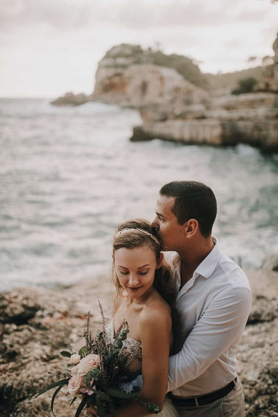 Romantisches Hochzeitsfoto am Strand mit verliebtem Paar und Blick auf die Küstenklippen in Tirol, Österreich.