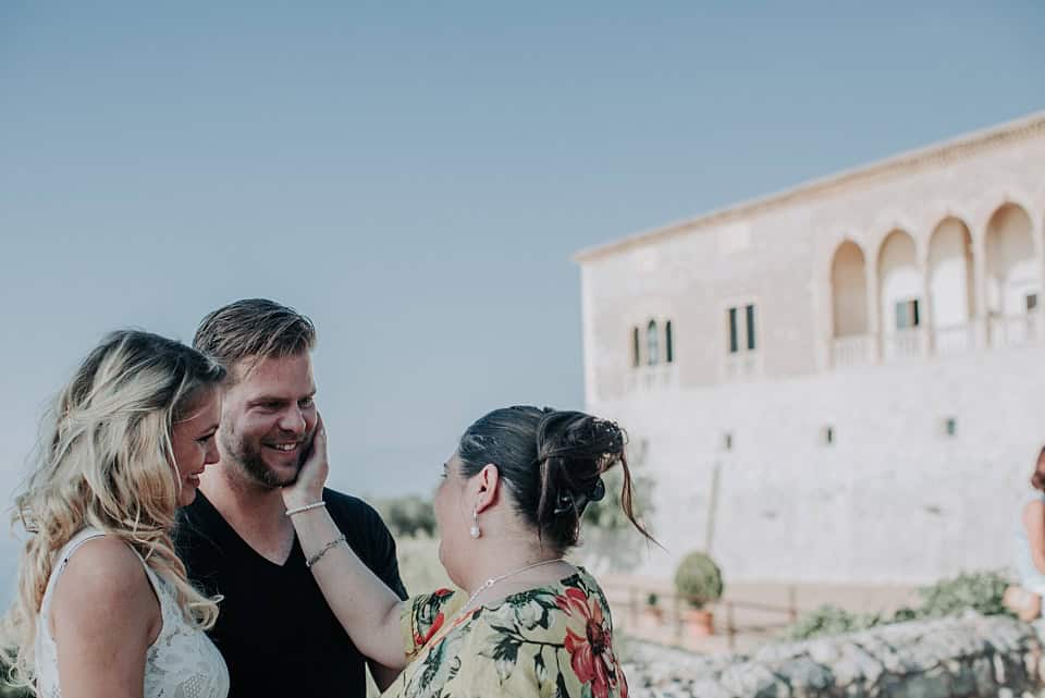 Fröhliche Hochzeitsgruppe am Meer vor antiker Steinfassade, gefühlvolle Momente, Mallorca, Hochzeitsfotografie, natürliche Stimmung.