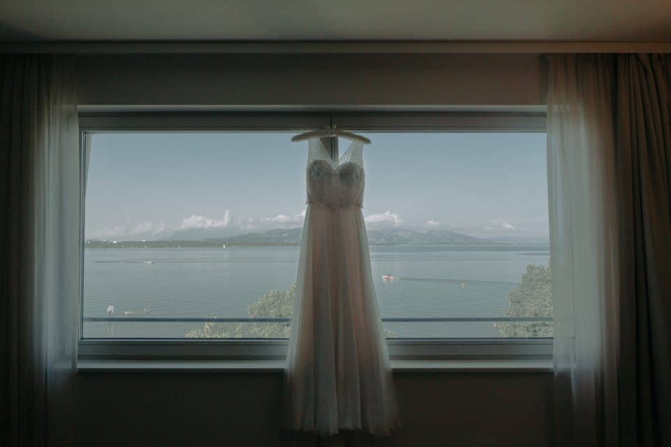 Elegantes Hochzeitskleid vor Fenster mit Seeblick in Tirol, Österreich.