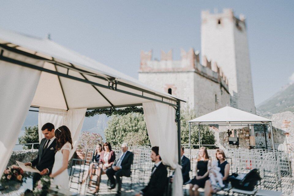 Hochzeit im Freien vor einer mittelalterlichen Burg, Brautpaar im Fokus, Gäste im Hintergrund, festliche Atmosphäre, Tirol Österreich.