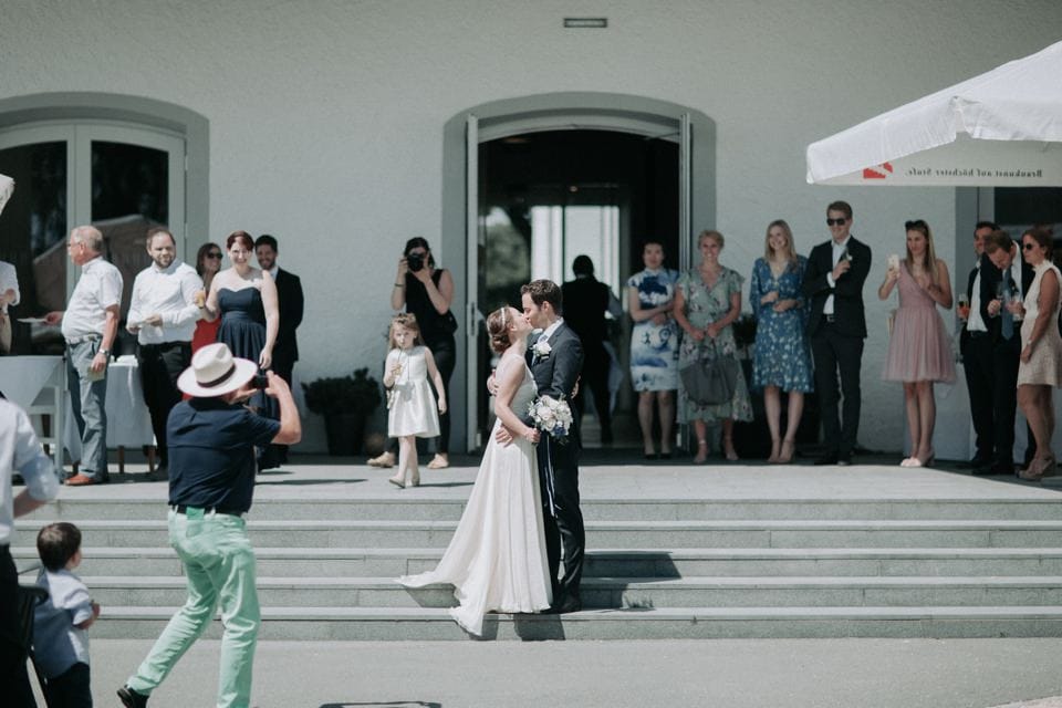 Hochzeit auf den Stufen vor einem weißen Gebäude mit Gästen im Hintergrund, romantisches Brautpaar im Fokus.
