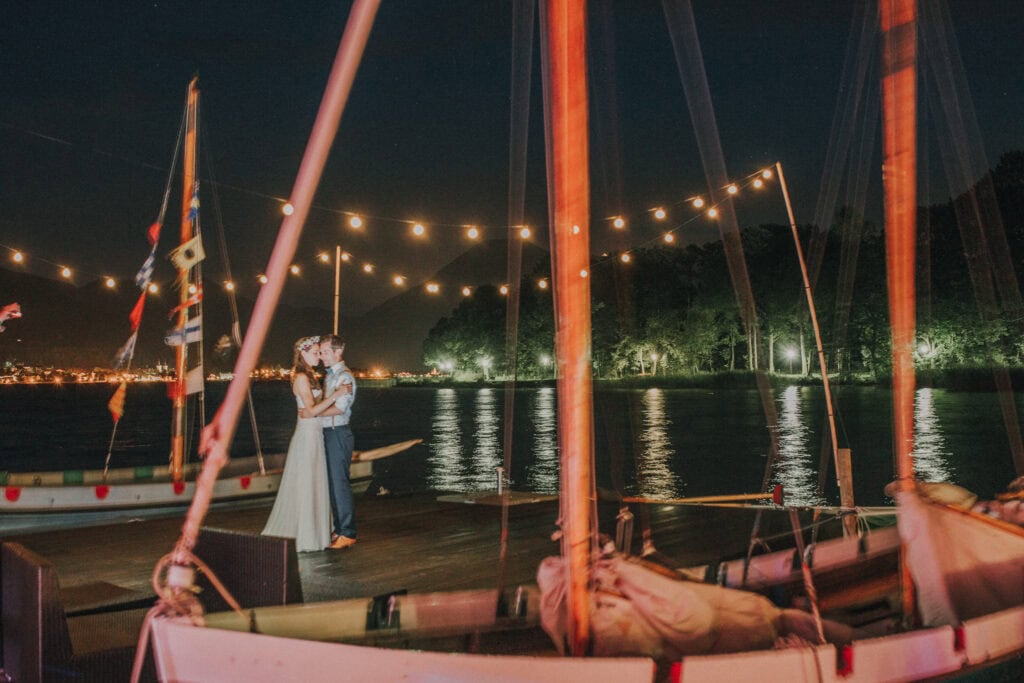 Romantisches Hochzeitspaar bei Nacht auf einem Boot am See, mit festlicher Beleuchtung im Hintergrund.