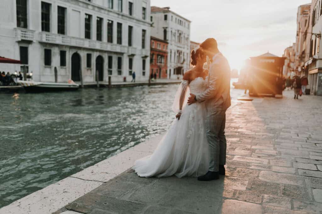Romantisches Hochzeitsfoto bei Sonnenuntergang in Venedig, Braut und Bräutigam am Wasser, kreative Hochzeitsfotografie in Italien.
