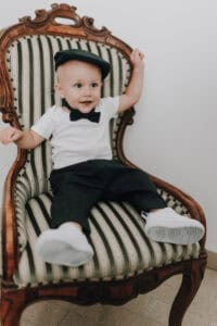 Schäbiger Junge im Smoking und kleiner Hut sitzt fröhlich auf antiker Polstersessel, Hochzeitsfotograf aus Tirol, Österreich.