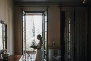 Hochzeitsbraut im Fensterlicht mit Blumen am Balkon, indoor und vintage Atmosphäre.