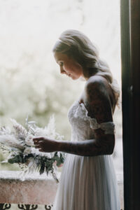 Elegante Braut mit Tattoo im Hochzeitskleid hält Blumenstrauß vor Fenster bei Tageslicht.