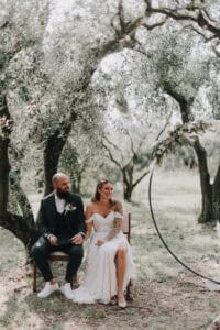 Natürliche Hochzeitsaufnahme im Olivenhain, Brautpaar sitzt auf Holzstühlen, romantische Atmosphäre, professionelle Hochzeitsfotografie Tirol Österreich international.