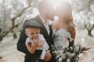 Romantisches Hochzeitsfoto mit Kind, Brautpaar mit Sonnenlicht, Natur im Hintergrund.