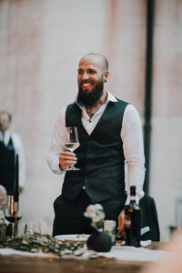 Fröhlicher Bräutigam beim Hochzeitsessen mit Wein in hand, stilvoll gekleidet in Tirol, Österreich.