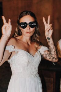 Lustige Braut mit Herz-Sonnenbrille und Peace-Zeichen bei Hochzeitsfeier in Tirol.