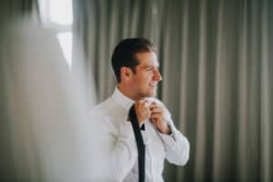 Bräutigam bindet Krawatte. Vorbereitung auf Hochzeit in Irland. Elegantes Outfit, helles Interieur.