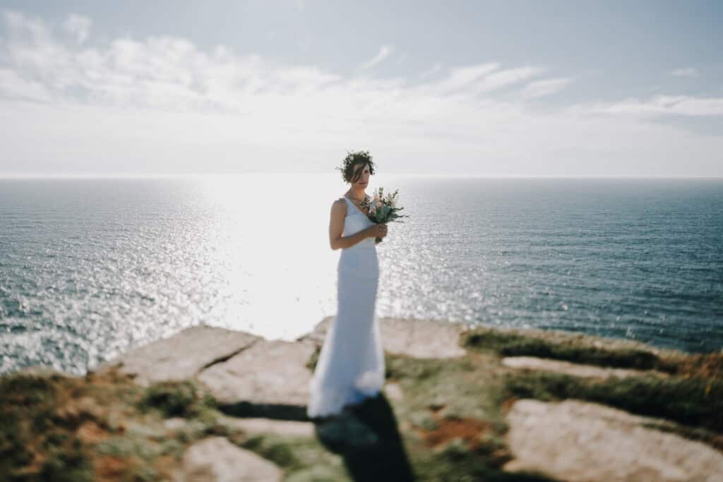 Braut mit Blumenkranz und Brautstrauß auf Klippe mit Meerblick. Hochzeitsfotograf Irland.
