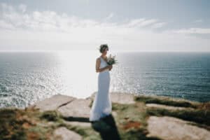 Braut mit Blumenkranz und Brautstrauß auf Klippe mit Meerblick. Hochzeitsfotograf Irland.