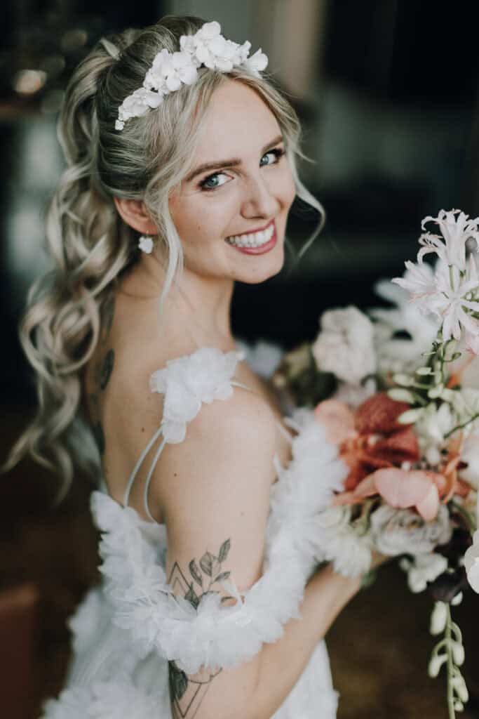 Braut mit Blumenstrauß und Blumenschmuck im Haar lächelt in die Kamera.