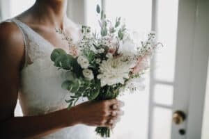 Braut hält weißen Blumenstrauß mit Eukalyptus vor weißem Kleid. Zarte Hochzeitsdetails.