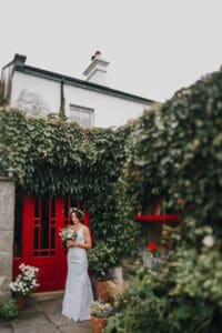 Braut mit Blumenstrauß vor roter Tür und grün bewachsenem irischen Haus. Hochzeitsfotograf Irland.