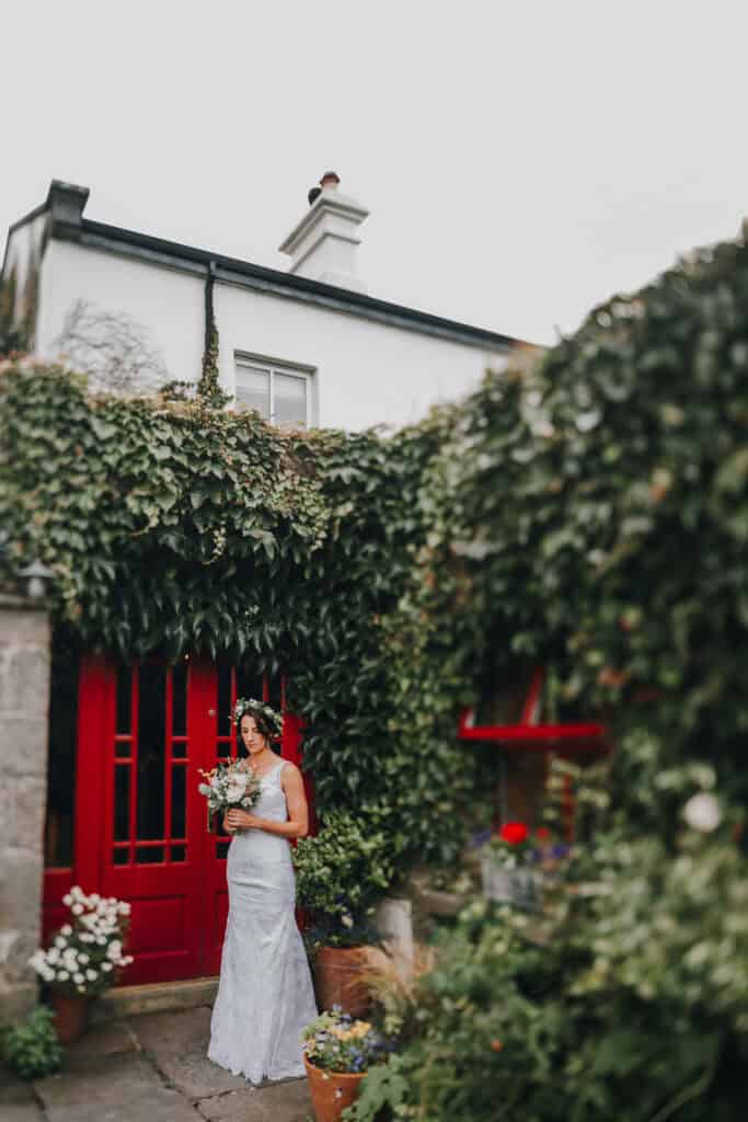 Braut mit Blumenstrauß vor roter Tür und grün bewachsenem irischen Haus. 