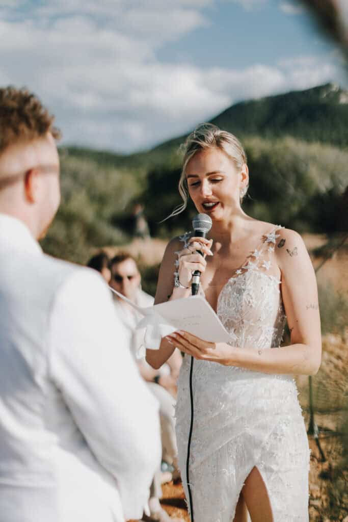 Braut hält Rede bei Ibiza Hochzeit. Elegantes Kleid, Mikrofon. Hügelige Landschaft im Hintergrund.
