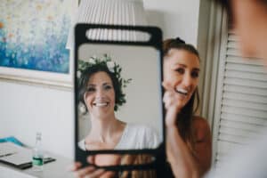 Braut blickt in Spiegel, gehalten von Brautjungfer. Blumenkranz im Haar, glücklich. Hochzeitsfotograf Irland.