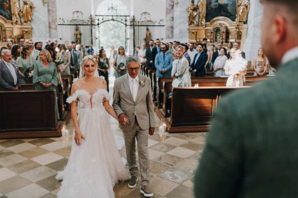 Braut mit Vater schreitet zum Altar in Kirche, umgeben von Hochzeitsgästen.