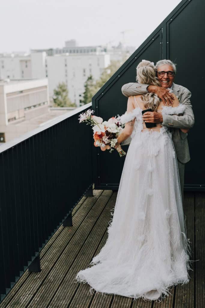 Braut umarmt Vater auf Balkon. Hochzeitsfotograf München. Zartes Brautkleid mit Federn.