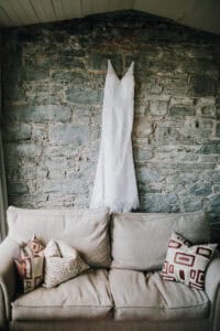 Weißes Brautkleid hängt vor einer Steinmauer über einem beigen Sofa mit gemusterten Kissen.