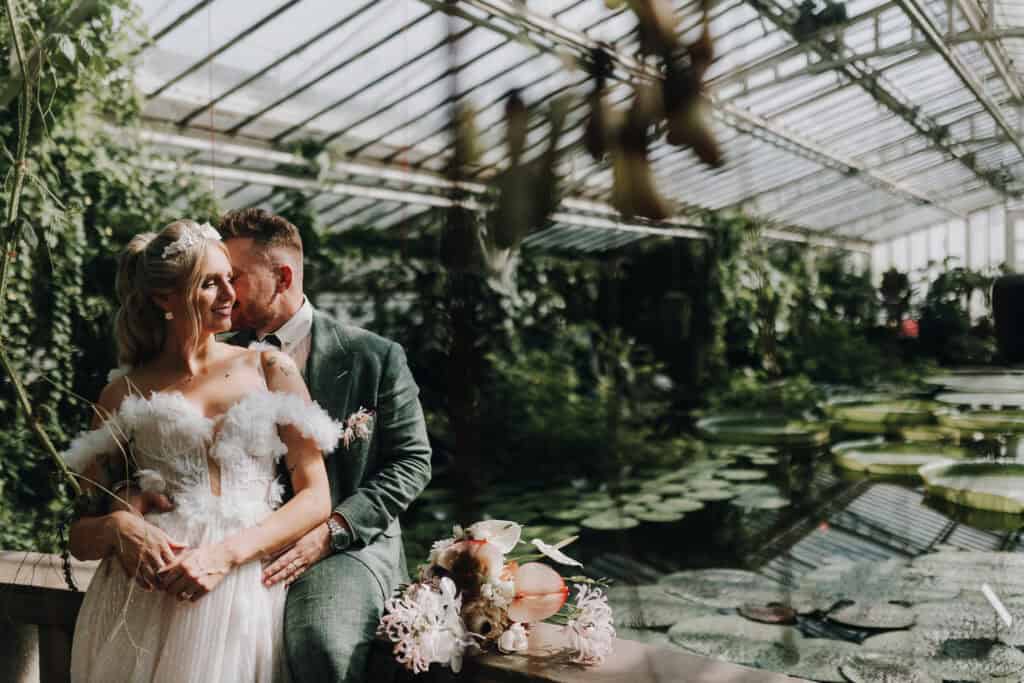 Brautpaar umarmt sich im Gewächshaus mit Teich. Hochzeitsfotograf München.