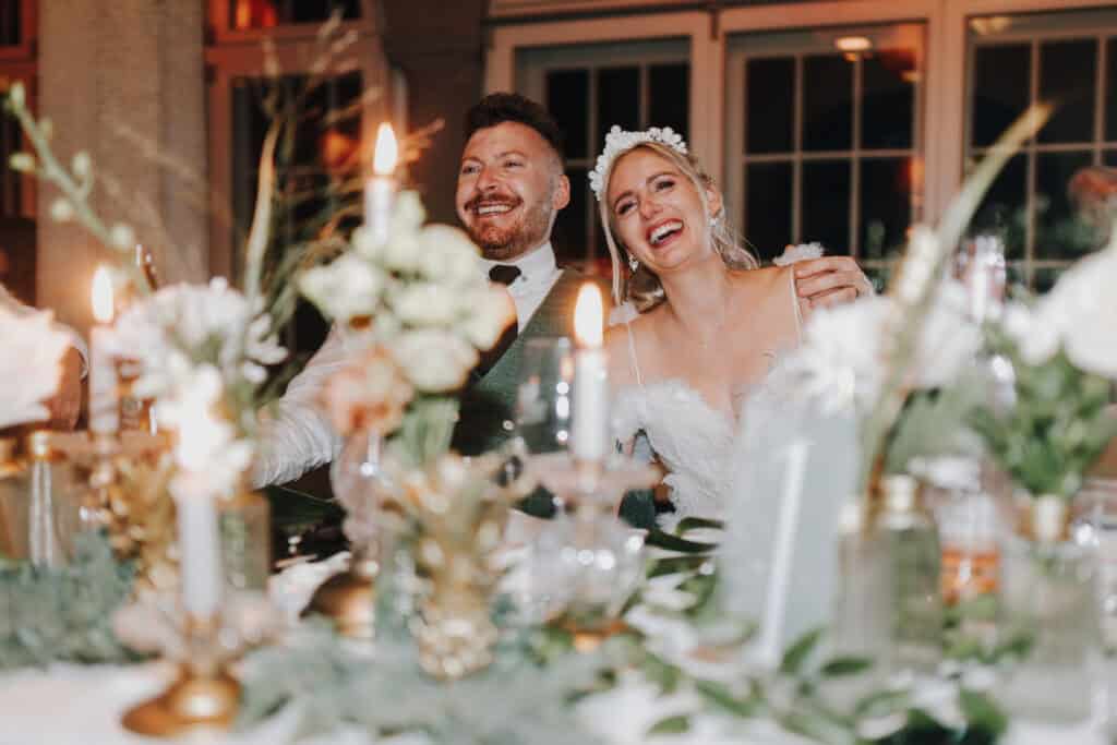 Fröhliches Brautpaar lacht am Hochzeitstisch mit Kerzen und Blumenschmuck. Hochzeit München.
