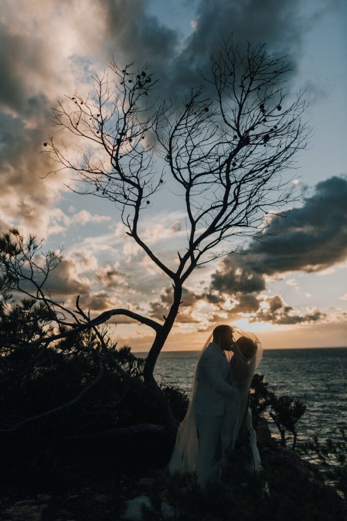 Brautpaar unter Schleier vor Ibiza-Küstenkulisse bei Sonnenuntergang. Romantische Hochzeit auf Ibiza.