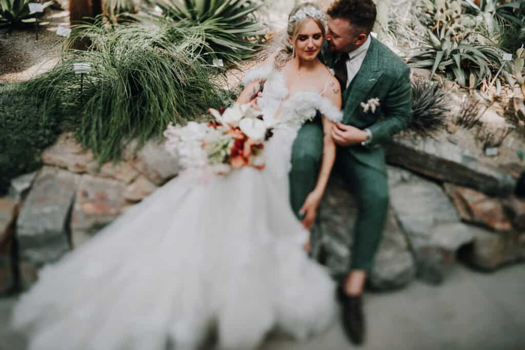 Brautpaar kuschelt in München, sie im weißen Kleid mit Blumenstrauß, er im grünen Anzug. Hochzeitsfotograf München.