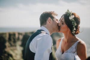 Brautpaar küsst sich innig vor irischer Küstenlandschaft. Braut mit Blumenkranz. Hochzeitsfotograf Irland.