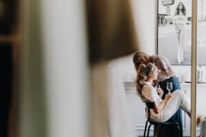 Brautstyling vor Hochzeit in München: Stylistin frisiert Braut mit Sektglas. Schwarz-Weiß-Foto an Wand.