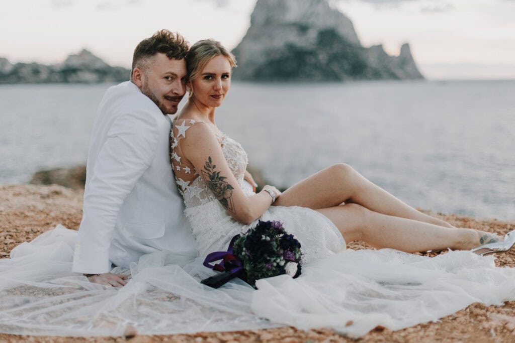 Brautpaar sitzt am Strand von Ibiza. Braut mit Tattoo und Bräutigam im weißen Anzug.