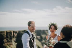 Lachendes Brautpaar in Irland mit Blumenkranz und Meerblick. Fröhliche Hochzeit im Freien.
