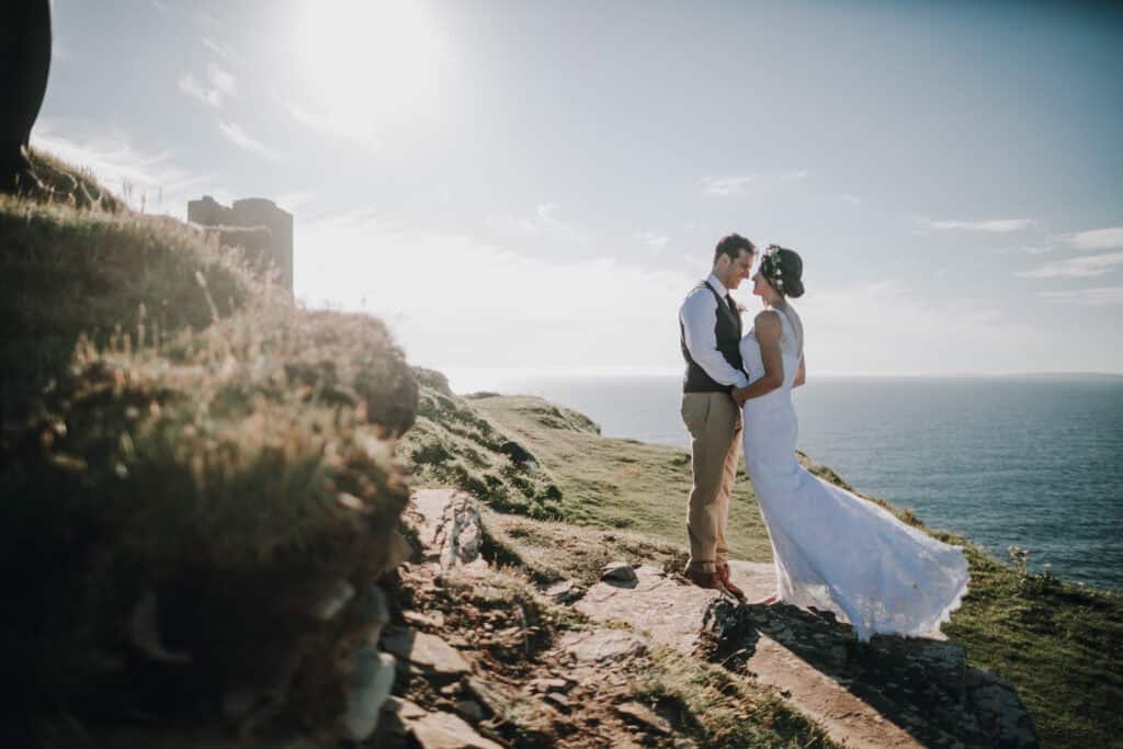 Brautpaar umarmt sich an irischer Küste. Ruine im Hintergrund. Sonnenlicht. Romantische Hochzeit in Irland.