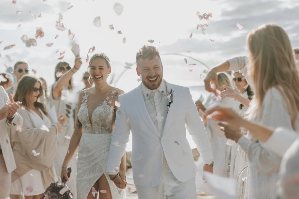 Brautpaar strahlt unter Blütenregen auf Ibiza. Glückliche Hochzeit, weiße Kleidung.