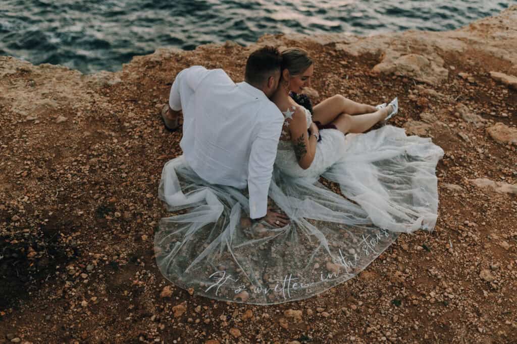 Brautpaar kuschelt auf Felsen mit Meerblick, Schleier mit Text "It's written in the stars".