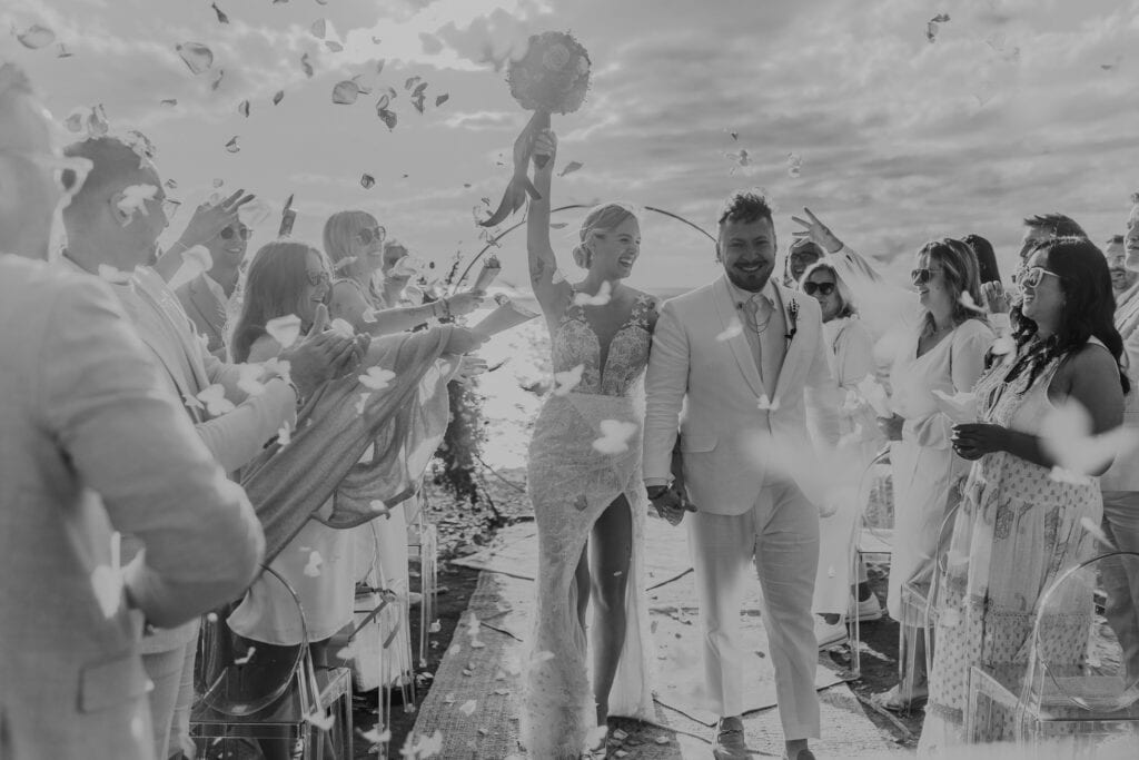 Brautpaar feiert Hochzeit auf Ibiza, umgeben von Gästen und Blütenblättern. Glückliche Momente.