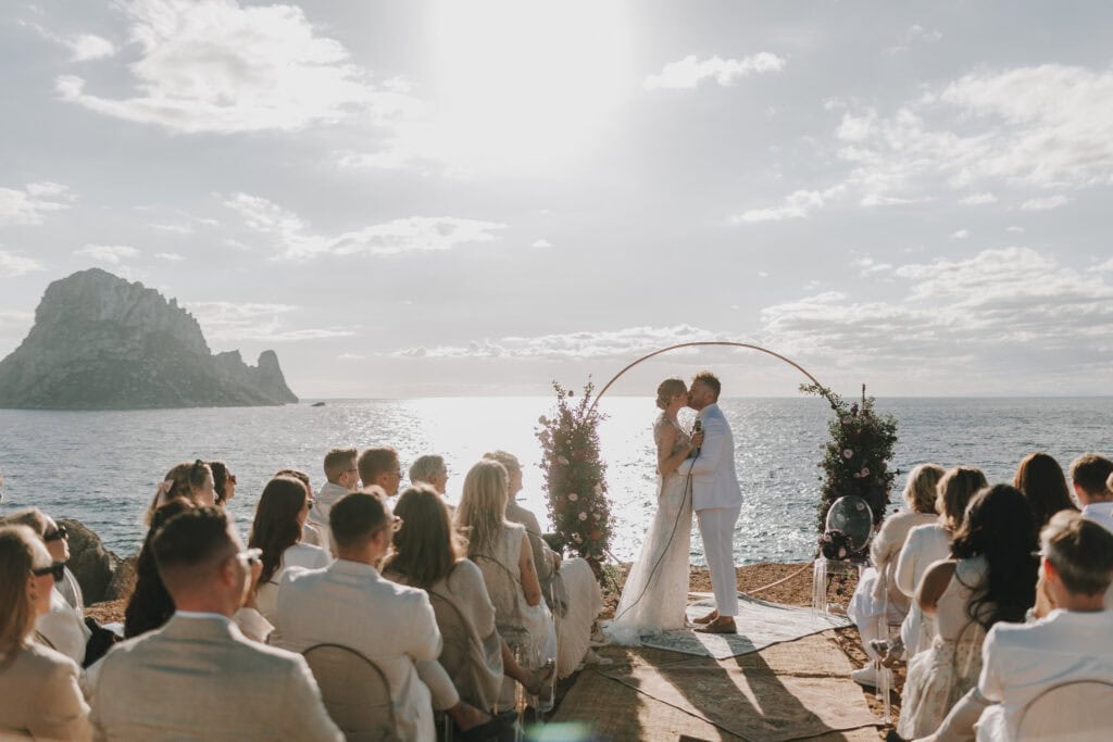 Brautpaar küsst sich bei Hochzeit auf Ibiza vor Es Vedrà. Gäste in Weiß.