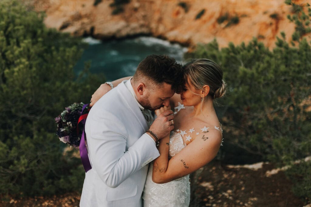Bräutigam küsst Hand der Braut vor Küstenkulisse. Romantische Hochzeit auf Ibiza.