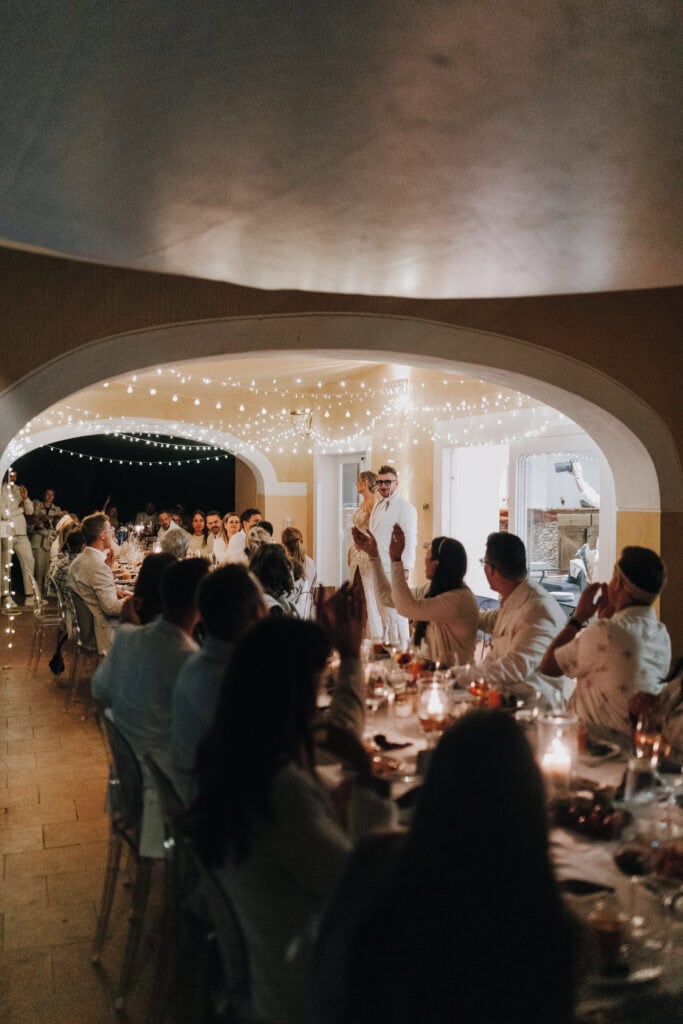 Brautpaar hält Rede bei Hochzeit auf Ibiza, Gäste applaudieren. Lichterketten schmücken den Raum.
