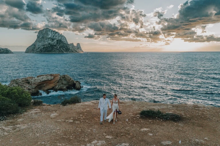 Brautpaar spaziert am Strand von Ibiza bei Sonnenuntergang mit Blick auf Es Vedrà. Romantische Hochzeit.