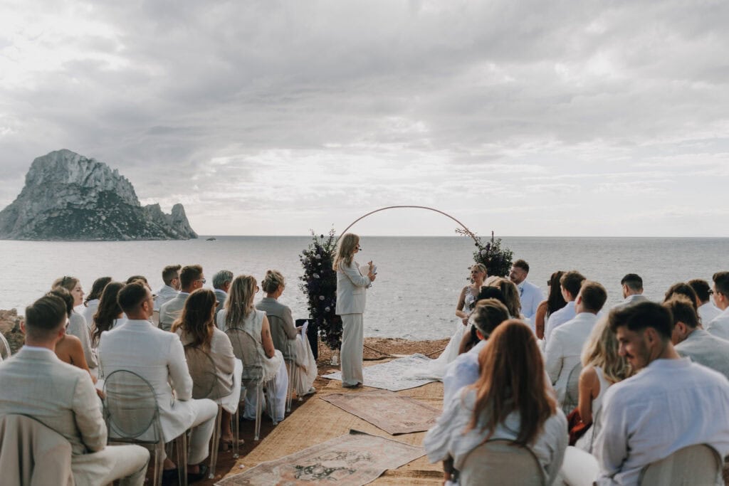 Hochzeit auf Ibiza: Brautpaar unter Bogen, Gäste in Weiß, Blick auf Felseninsel.