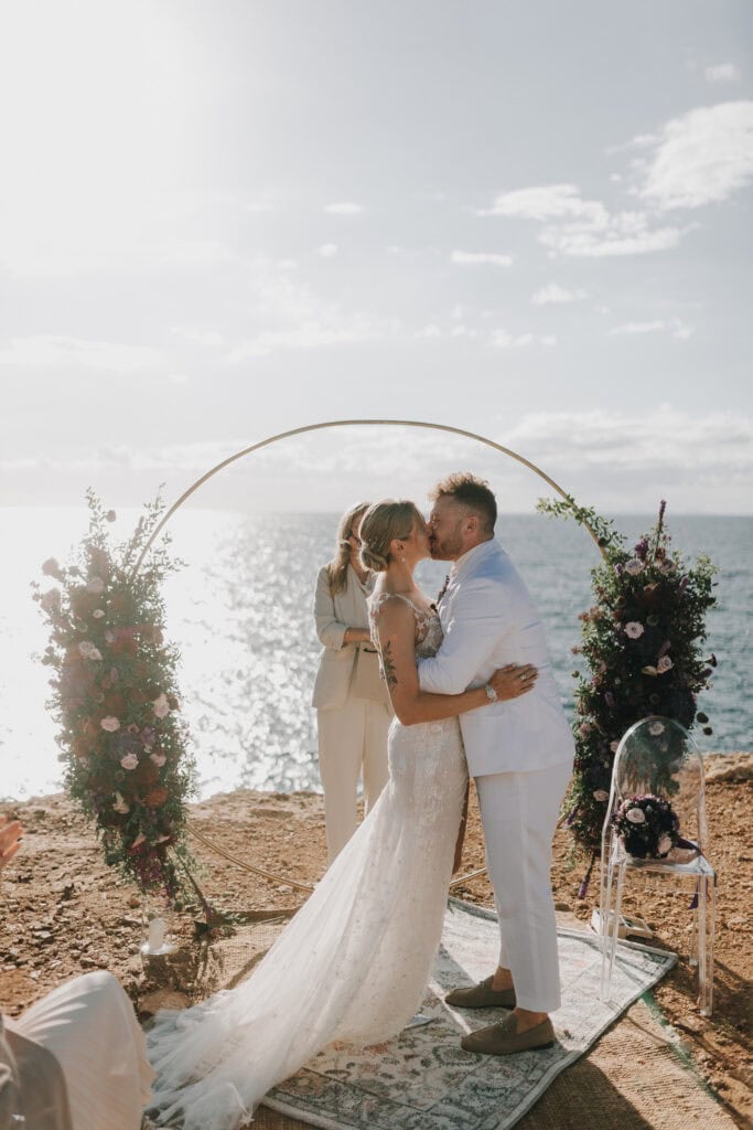 Brautpaar küsst sich unter Hochzeitsbogen auf Ibiza mit Meerblick. Hochzeitsfotograf Ibiza.
