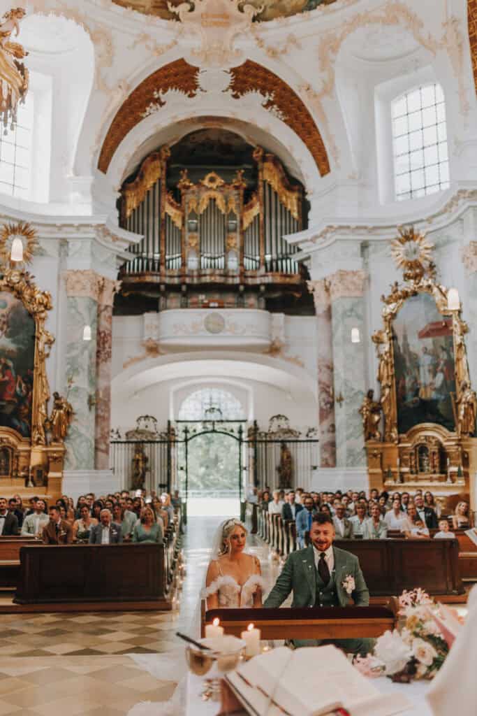 Brautpaar in Kirche mit Gästen. Barocke Hochzeitslocation in München.