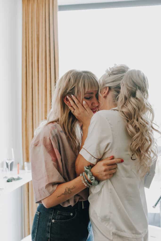 Zwei blonde Frauen umarmen sich liebevoll vor einem Fenster. Zärtlicher Moment, Hochzeitsfotograf München.