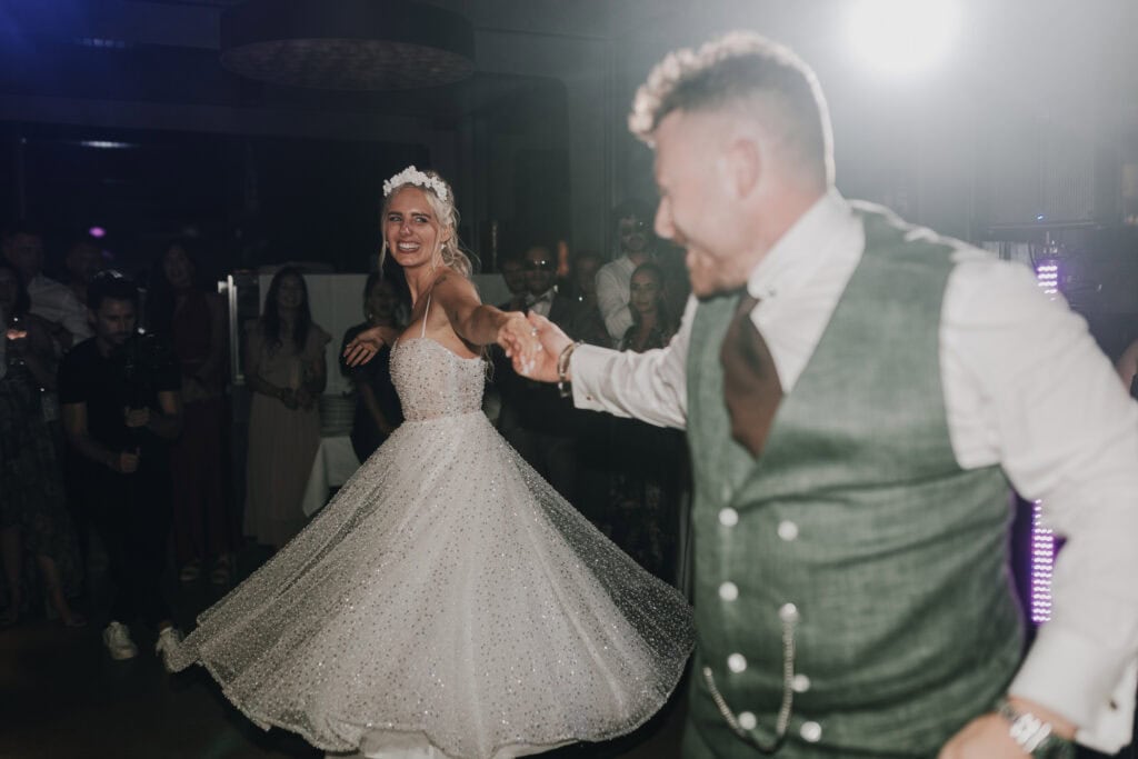 Fröhliches Brautpaar tanzt auf der Hochzeit in München. Die Braut trägt ein glitzerndes Kleid, der Bräutigam eine grüne Weste.
