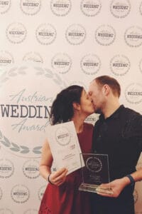 Paar küsst sich vor "Austrian Wedding Award"-Hintergrund, hält Urkunde und Preis. Auszeichnung für Hochzeitsfotografie.
