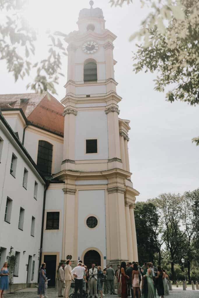 Hochzeitsgesellschaft vor barocker Kirche in München. Brautpaar mit Gästen im Freien.