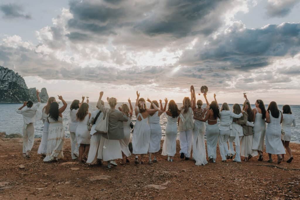 Gruppe Frauen in Weiß jubeln am Strand von Ibiza unter bewölktem Himmel.